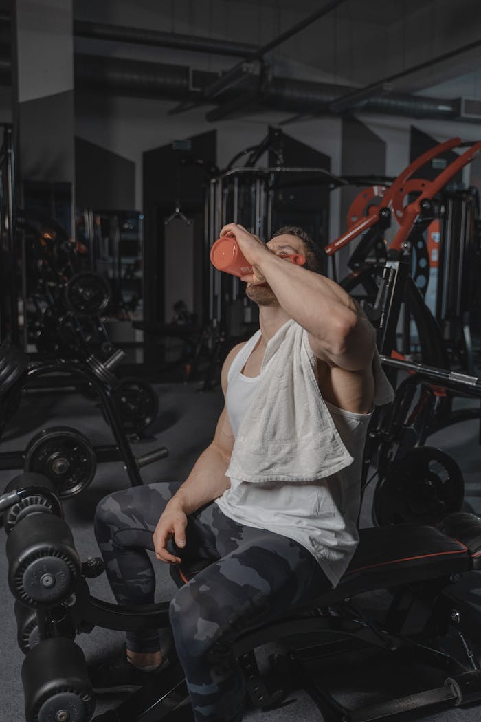 A muscular man with a towel rests and drinks water after an intense workout in a modern gym.
