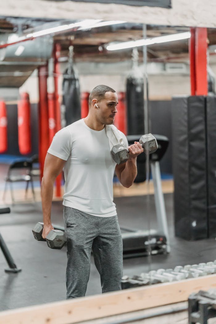 Young African American male athlete doing dumbbells curls while standing in front of mirror during fitness workout in modern gym